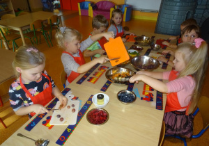 Grupa dzieci siedzi przy stole, w ręku trzymają noże, którymi kroją owoce na desce. Jedna z dziewczynek zrzuca pokrojone owoce z deski do miski.